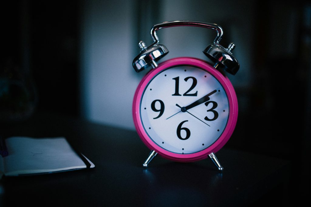 Image shows an alarm clock with two big silver bells on the top. The clock's surround is bright pink. It's face is white. It has 12, 3, 6 and 9 in large black numbers. The hands are pointing to ten past two. The background is dark so it might be night time.